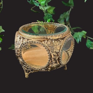May include: A gold-tone trinket box with a glass lid and intricate floral designs. The box has a round shape and sits on three legs.
