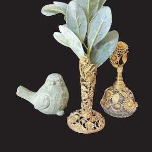 May include: A collection of decorative items on a black background. Includes a light green bird figurine, a gold-colored vase with sunflower detailing holding green leafy stems, and a gold-colored perfume bottle with a sunflower design.
