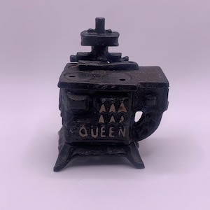 Puede incluir: Estufa de juguete de hierro fundido negro con la palabra "QUEEN" y tres triángulos en la parte delantera.