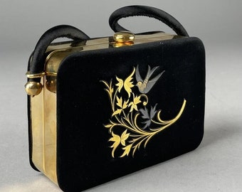 Puede incluir: Un bolso de mano vintage de terciopelo negro con detalles metálicos dorados. El bolso presenta un diseño decorativo de un pájaro y follaje en oro, gris y negro. Tiene un asa negra y un cierre de broche.