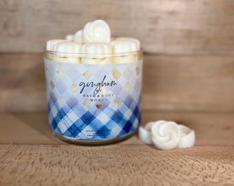 Può includere: Un barattolo trasparente di cera profumata con un motivo a quadri blu e bianco. Il barattolo è pieno di cere bianche a forma di fiore. Le parole "gingham" e "Bath & Body Works" sono stampate sul barattolo.
