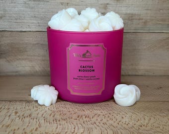 Può includere: Un portacandele rosa acceso pieno di cera profumata bianca a forma di rosa. L'etichetta recita "Cactus Blossom" con note di petali di fiori di cactus, agrumi freschi e cocco vanigliato. Due cere profumate sono fuori.