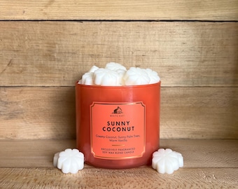 Può includere: Una candela arancione con il testo "Sunny Coconut" su un'etichetta. La candela è riempita con cere profumate bianche e si trova su una superficie di legno. Due cere profumate bianche sono accanto alla candela.