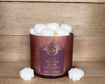 Może przedstawiać: Miedziany słoik na świecę wypełniony białymi woskami zapachowymi w kształcie kwiatów. Etykieta zawiera napis White Barn, Vanilla Bean, Madagascar Vanilla, Homemade Marshmallow, Nutmeg, Exclusively Fragranced Soy Wax Blend Candle. Dwa woski leżą z przodu.