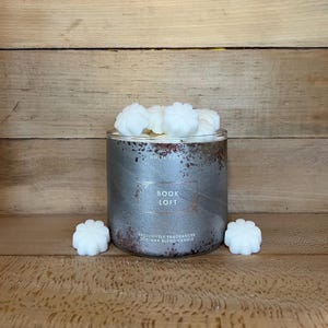 May include: A silver-colored candle jar with the words "BOOK LOFT" printed on the front. The jar is filled with white wax melts and sits on a wooden surface. Two white wax melts are placed near the jar.