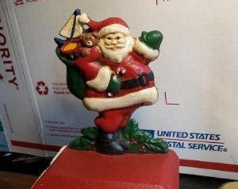 May include: A red and white cast iron Santa Claus stocking holder with a green bag and a teddy bear. The Santa Claus is holding the bag and the teddy bear in his arms. The stocking holder is on a red base with a hook on the bottom.