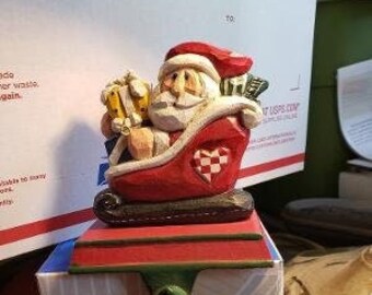May include: A red and white stocking holder featuring a Santa Claus figurine holding a gift box. The Santa Claus is riding in a red sleigh with a heart-shaped design. The stocking holder is on a white box with a geometric pattern.