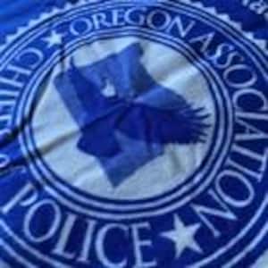 May include: A blue and white t-shirt featuring the text "OREGON ASSOCIATION POLICE CHIEFS" in a circular design. The design includes a blue eagle and a map of Oregon.