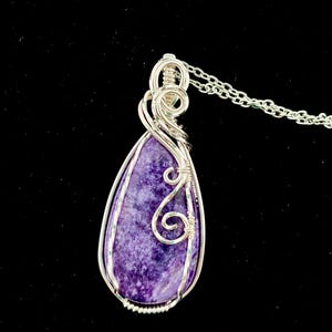 Puede incluir: Un colgante de charoíta morada en forma de lágrima, envuelto en alambre de plata con intrincadas espirales. El colgante cuelga de una cadena de plata y presenta una piedra pulida con patrones veteados en blanco y violeta oscuro.