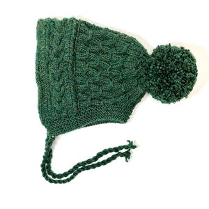 May include: A dark green knitted beanie with a pom pom on top and a long, tied string. The beanie has a cable knit pattern.