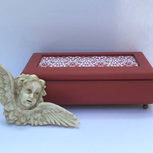 May include: A red wooden box with a white and red patterned lid. The box is closed and has a small angel figurine in front of it.