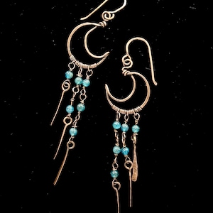 May include: Silver-toned crescent moon earrings with dangling strands of blue beads and elongated metal accents. The earrings have a unique, handcrafted design, perfect for adding a touch of celestial charm to any outfit.