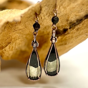 May include: A pair of handcrafted earrings with a copper wire frame. Each earring features a teardrop-shaped, dark green stone with a silver-colored section. Small black beads sit above the stone. The earrings hang from copper hooks.