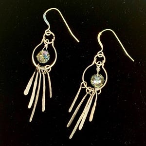 Puede incluir: Un par de pendientes colgantes plateados. Cada pendiente presenta un marco de alambre en forma de lágrima con una cuenta iridiscente facetada y varias barras plateadas colgantes. Los pendientes tienen un cierre de gancho.