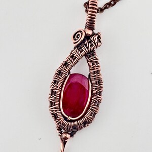 May include: A copper wire pendant necklace with a teardrop design. The pendant features a vivid, oval-cut, red gemstone. The intricate wirework and the stone's colour create a unique piece of jewellery. The necklace is suspended from a copper chain.