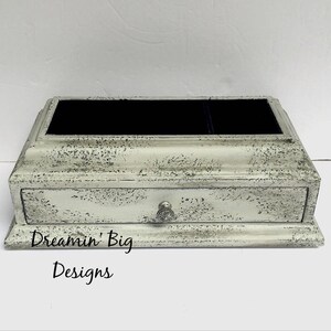 May include: A white wooden jewelry box with a distressed finish. The box has a drawer and a top compartment with a black velvet lining. The box is labeled "Dreamin' Big Designs".