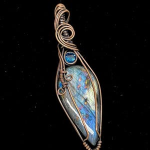 Puede incluir: Un colgante artesanal con una piedra de labradorita en forma de pera con tonos iridiscentes azules y dorados, envuelta en alambre de cobre. Una pequeña gema azul está engastada cerca de la parte superior, y el trabajo de alambre incluye espirales decorativas.