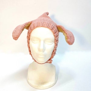 Puede incluir: Un gorro de punto rosa con orejas caídas, que se asemejan a las orejas de un perro.