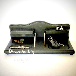 Puede incluir: Un organizador de madera negro con múltiples compartimentos para llaves, monedas y otros objetos pequeños. El organizador tiene una parte superior curva y un logotipo "Dreamin' Big Designs".