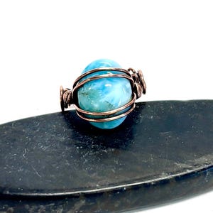 Puede incluir: Un anillo artesanal con una piedra preciosa azul claro, envuelta en alambre de cobre. La piedra tiene un aspecto veteado con tonos azules y blancos. El anillo está sobre una superficie oscura y texturizada, creando un contraste.