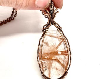 May include: A copper wire wrapped pendant featuring a clear quartz crystal with brown inclusions. The pendant is attached to a copper chain.