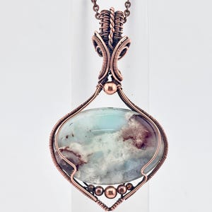 May include: A handcrafted copper wire-wrapped pendant necklace. The pendant features an oval-shaped stone with a mix of blue, white, and brown hues. The copper wire is intricately woven around the stone and forms the necklace's chain.