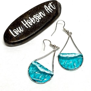 May include: Teardrop-shaped earrings with a silver frame and a turquoise resin design resembling ocean waves. The earrings have a hook closure. A dark stone with the text "Lou Hobson Art" is in the background.