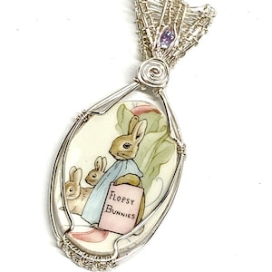 May include: Silver pendant featuring a hand-painted illustration of rabbits from the "Flopsy Bunnies" story. The pendant has an oval shape with a wire-wrapped design and a small purple gemstone at the top. The text "Flopsy Bunnies" is visible.