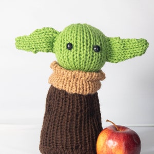 Puede incluir: Un juguete de peluche verde tejido con orejas grandes y ropa de punto marr&oacute;n. El juguete est&aacute; de pie junto a una manzana roja.