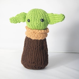 Puede incluir: Un juguete de peluche verde tejido con orejas grandes y ropa de punto marr&oacute;n. El juguete tiene ojos de bot&oacute;n negros.