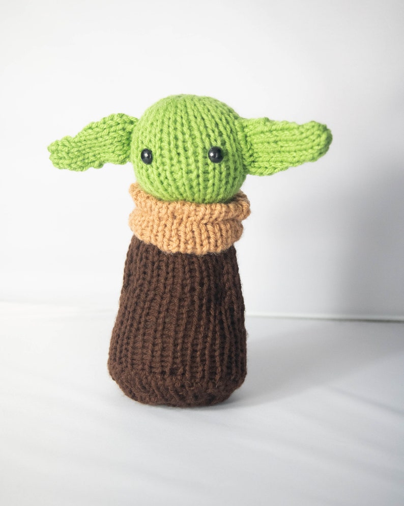 Puede incluir: Un juguete de peluche verde tejido con orejas grandes y ropa de punto marr&oacute;n. El juguete tiene ojos de bot&oacute;n negros.