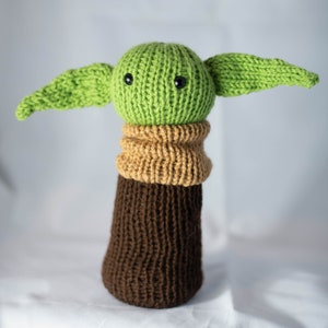 K&ouml;nnte beinhalten: Ein gr&uuml;nes Strickspielzeug mit gro&szlig;en Ohren, brauner und beiger Strickkleidung und schwarzen Augen. Das Spielzeug ist ein Charakter aus dem Star Wars-Franchise.