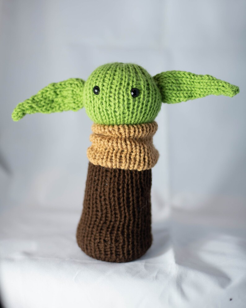 K&ouml;nnte beinhalten: Ein gr&uuml;nes Strickspielzeug mit gro&szlig;en Ohren, brauner und beiger Strickkleidung und schwarzen Augen. Das Spielzeug ist ein Charakter aus dem Star Wars-Franchise.