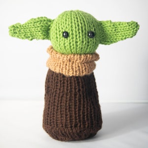Puede incluir: Un juguete de peluche verde y marr&oacute;n tejido con orejas grandes y ojos negros. El juguete es un personaje popular de la franquicia Star Wars.