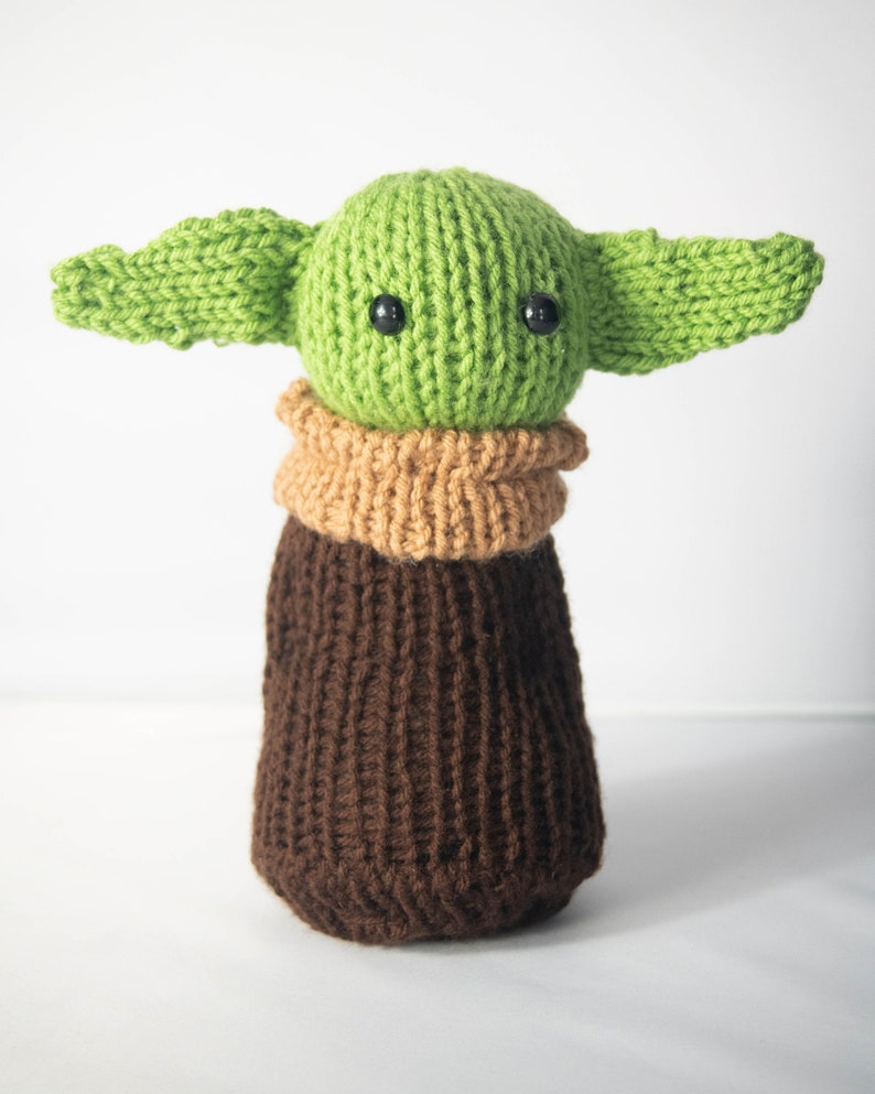 Puede incluir: Un juguete de peluche verde y marr&oacute;n tejido con orejas grandes y ojos negros. El juguete es un personaje popular de la franquicia Star Wars.