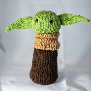 K&ouml;nnte beinhalten: Ein gestricktes gr&uuml;nes und braunes Pl&uuml;schtier mit gro&szlig;en Ohren und schwarzen Augen. Das Spielzeug ist in Form eines Charakters aus dem Star Wars-Franchise.