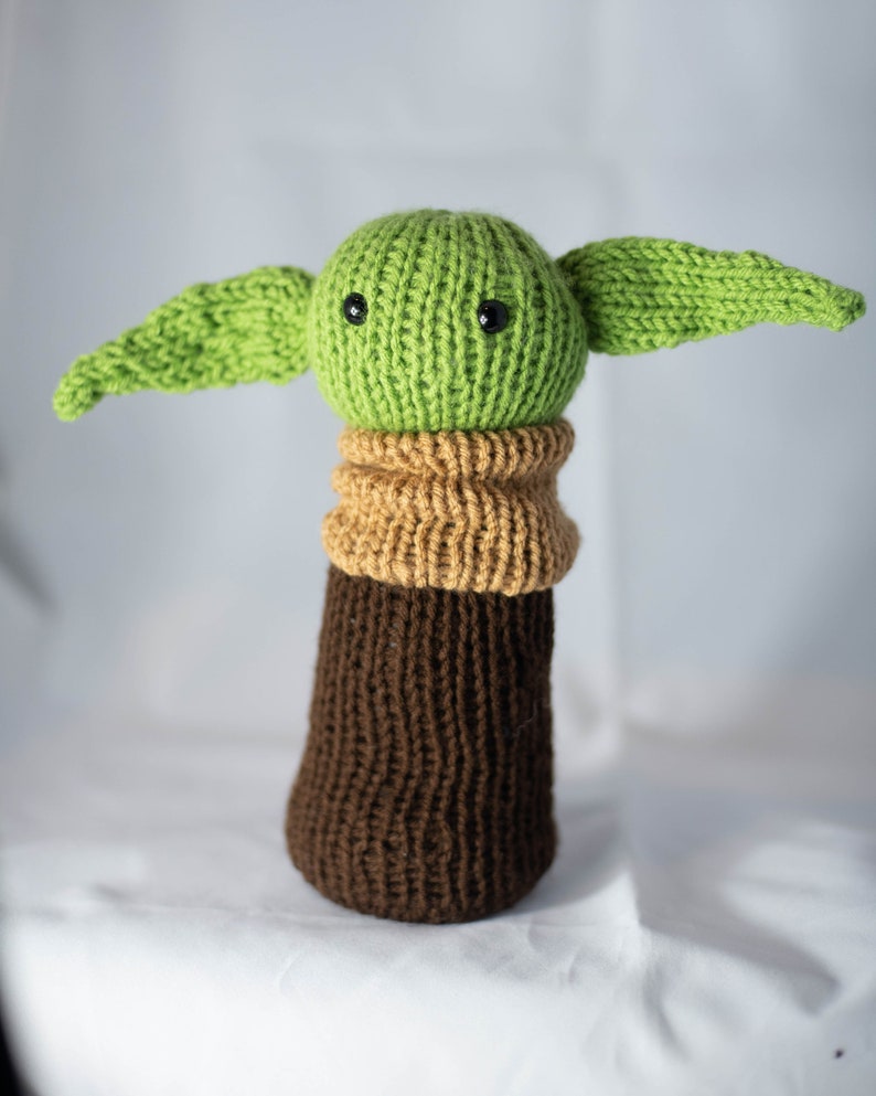 K&ouml;nnte beinhalten: Ein gestricktes gr&uuml;nes und braunes Pl&uuml;schtier mit gro&szlig;en Ohren und schwarzen Augen. Das Spielzeug ist in Form eines Charakters aus dem Star Wars-Franchise.