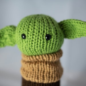 K&ouml;nnte beinhalten: Ein gr&uuml;nes, gestricktes Pl&uuml;schtier mit gro&szlig;en schwarzen Augen und brauner Strickkleidung. Das Spielzeug ist ein beliebter Charakter aus dem Star Wars-Franchise.