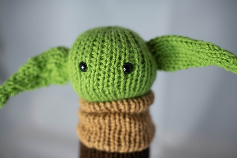 K&ouml;nnte beinhalten: Ein gr&uuml;nes, gestricktes Pl&uuml;schtier mit gro&szlig;en schwarzen Augen und brauner Strickkleidung. Das Spielzeug ist ein beliebter Charakter aus dem Star Wars-Franchise.