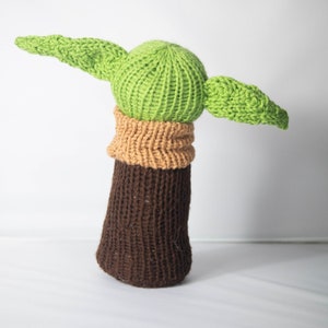 Puede incluir: Un juguete de peluche tejido con forma de personaje de la franquicia Star Wars. El juguete es verde con orejas grandes y un cuerpo marr&oacute;n.