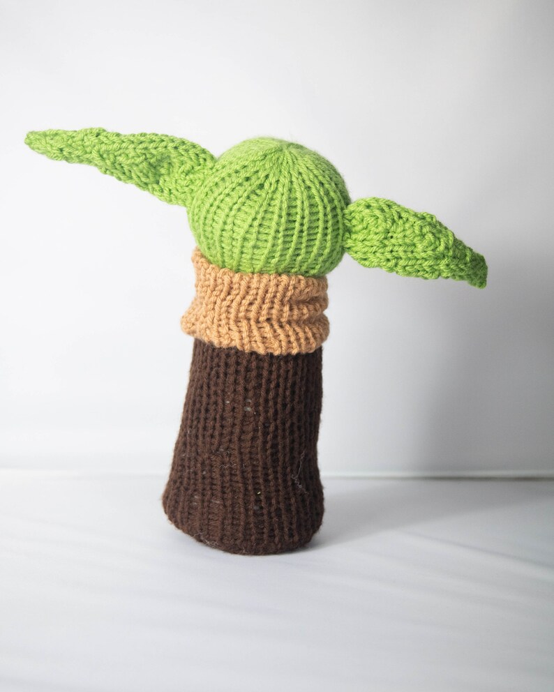 Puede incluir: Un juguete de peluche tejido con forma de personaje de la franquicia Star Wars. El juguete es verde con orejas grandes y un cuerpo marr&oacute;n.