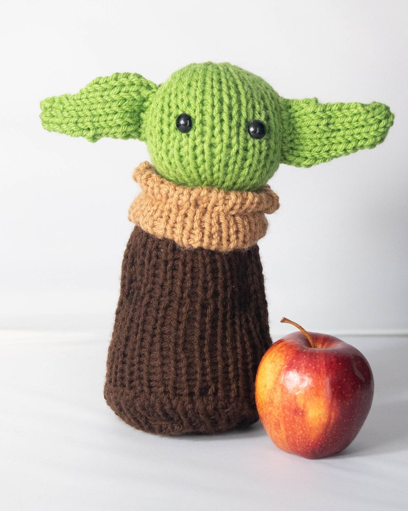 Puede incluir: Un juguete de peluche verde tejido con orejas grandes y ropa de punto marr&oacute;n. El juguete est&aacute; de pie junto a una manzana roja.