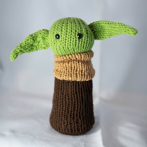 K&ouml;nnte beinhalten: Ein gr&uuml;nes Strickspielzeug mit gro&szlig;en Ohren und schwarzen Augen, das einen braunen und beigefarbenen Strickpullover tr&auml;gt.