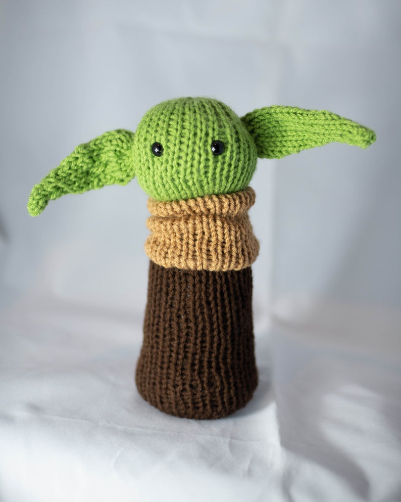 K&ouml;nnte beinhalten: Ein gr&uuml;nes Strickspielzeug mit gro&szlig;en Ohren und schwarzen Augen, das einen braunen und beigefarbenen Strickpullover tr&auml;gt.