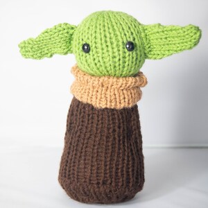 Puede incluir: Un juguete de peluche verde y marr&oacute;n tejido con orejas grandes y ojos negros. El juguete tiene la forma de un personaje de la franquicia Star Wars.