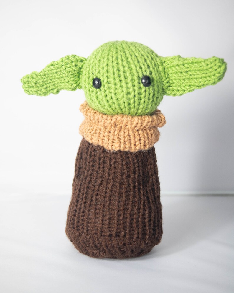 Puede incluir: Un juguete de peluche verde y marr&oacute;n tejido con orejas grandes y ojos negros. El juguete tiene la forma de un personaje de la franquicia Star Wars.