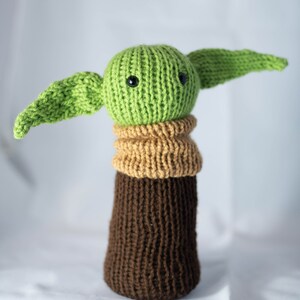 K&ouml;nnte beinhalten: Ein gestricktes gr&uuml;nes und braunes Pl&uuml;schtier mit gro&szlig;en Ohren und schwarzen Augen. Das Spielzeug ist ein beliebter Charakter aus dem Star Wars-Franchise.