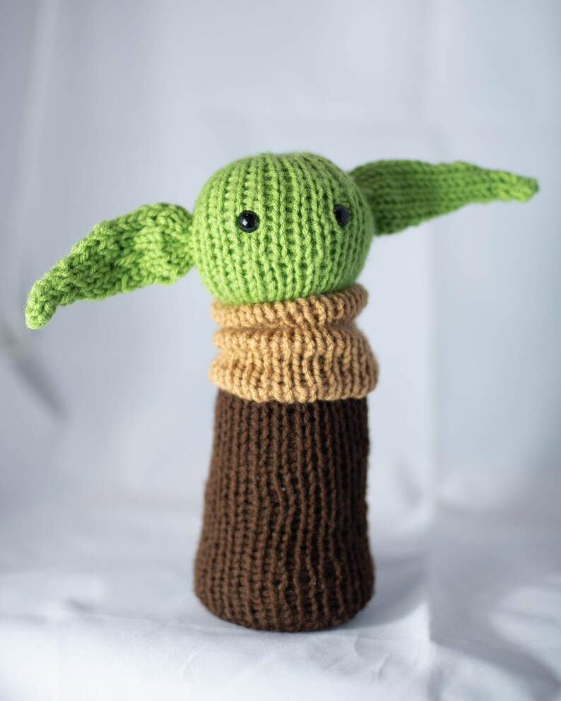 K&ouml;nnte beinhalten: Ein gestricktes gr&uuml;nes und braunes Pl&uuml;schtier mit gro&szlig;en Ohren und schwarzen Augen. Das Spielzeug ist ein beliebter Charakter aus dem Star Wars-Franchise.