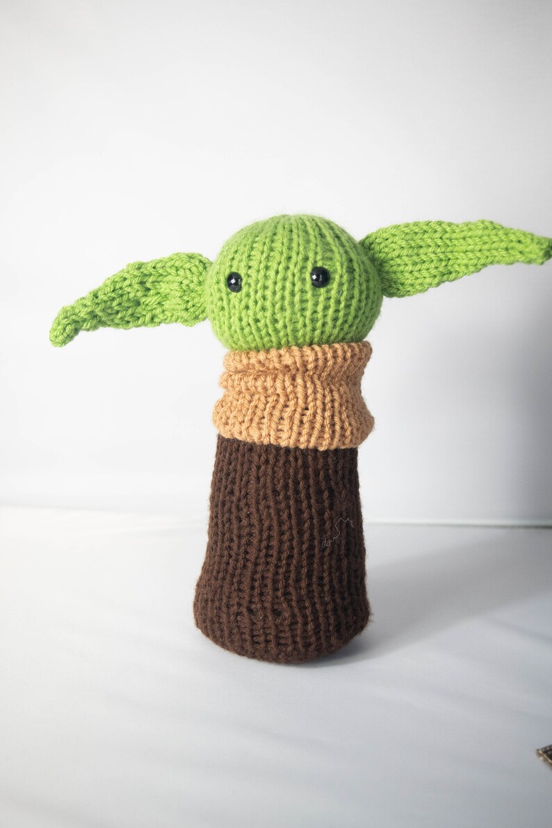 K&ouml;nnte beinhalten: Ein gr&uuml;nes, gestricktes Pl&uuml;schtier mit gro&szlig;en Ohren und braunen, gestrickten Beinen. Das Spielzeug hat schwarze Knopfaugen und einen gestrickten braunen Kragen.