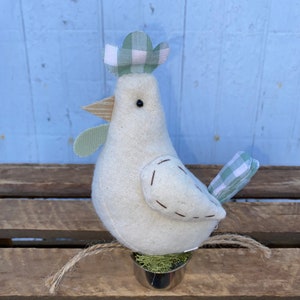 Puede incluir: Un pollo de tela blanca con un sombrero a cuadros verde y blanco, un pico marr&oacute;n y ojos negros. El pollo est&aacute; sentado en una maceta peque&ntilde;a con musgo verde.